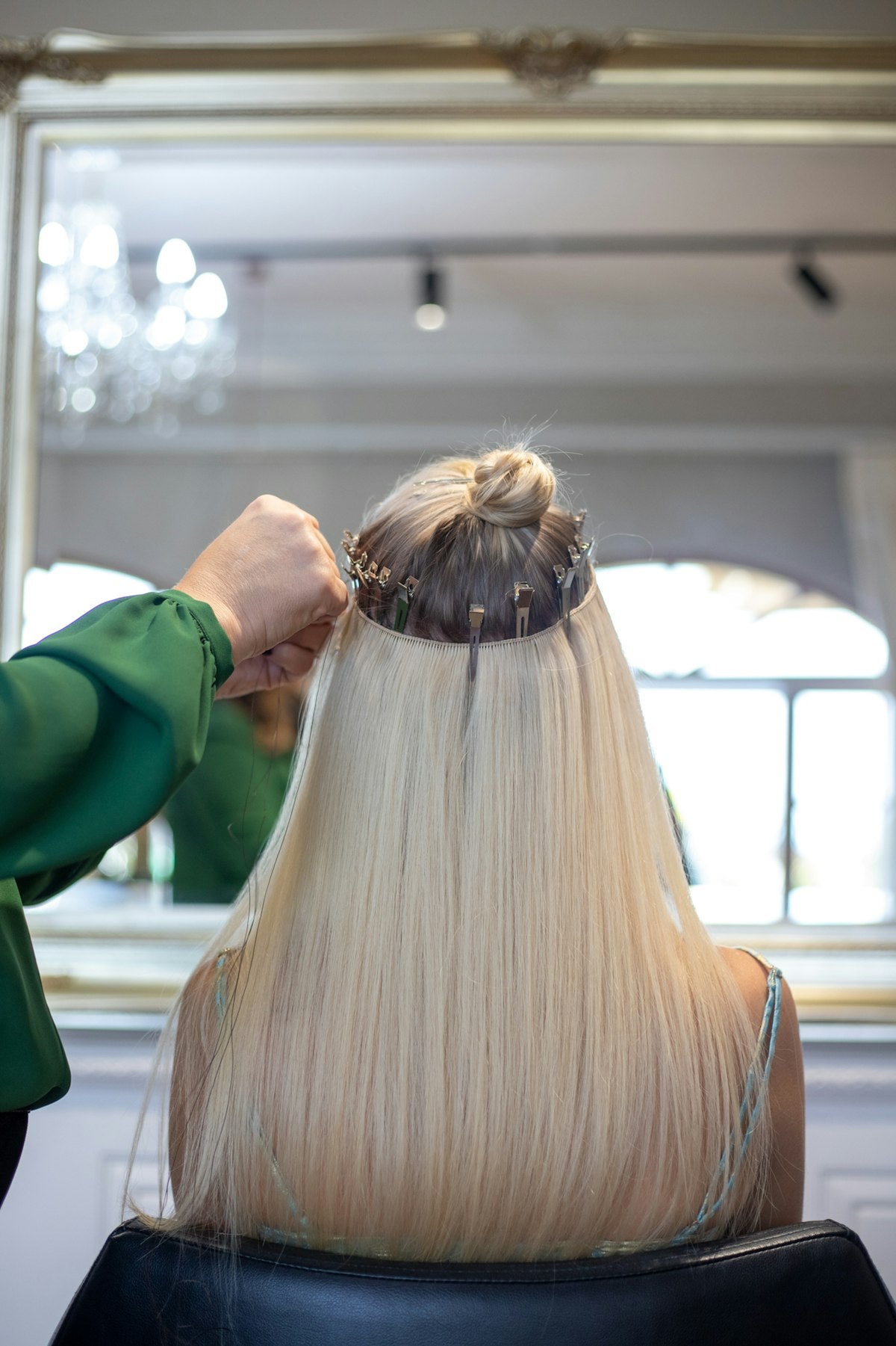 A woman seen from behind in a salon chair while a stylist fits long straight blonde extensions